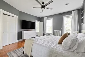 Quarto com cama, televisão, ventoinha no teto e janela com cortinas.