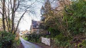 A narrow rural road flanked by dense foliage and a stone house with a white picket fence. - Family home with sea view: garden & beach nearby (Ploubazlanec)