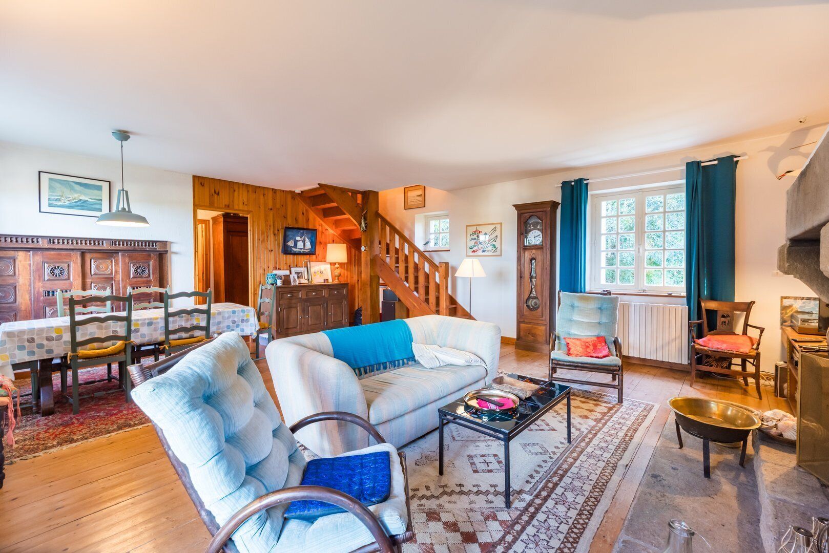 Una sala de estar acogedora con sofá, mesa de comedor y sillas, una escalera y un ventanal con cortinas.