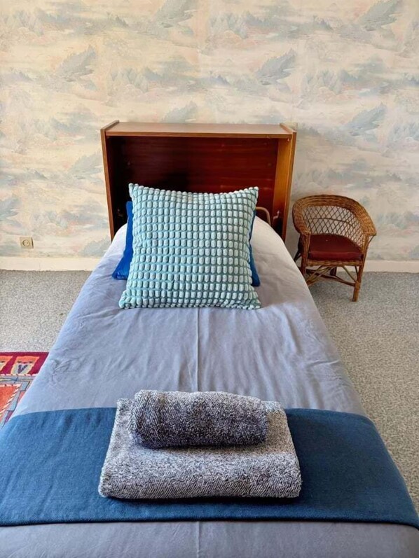 A bed with a wooden headboard, a blue and white patterned pillow, a grey blanket, and a folded grey towel.