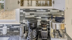 Ett kök med öppna skåp där vinglas, muggar och svarta förvaringsbehållare står framme. På bänkskivan finns en kaffebryggare och en vattenkokare.