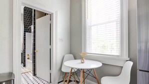 Een kleine kamer met een zwart-wit geruite vloer, een witte tafel met twee stoelen, een raam met jaloezieën en een deur die uitkomt op een betegeld gebied.