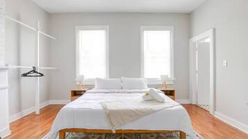 A bedroom with a bed, two windows, a wooden floor, and a hanging coat rack.