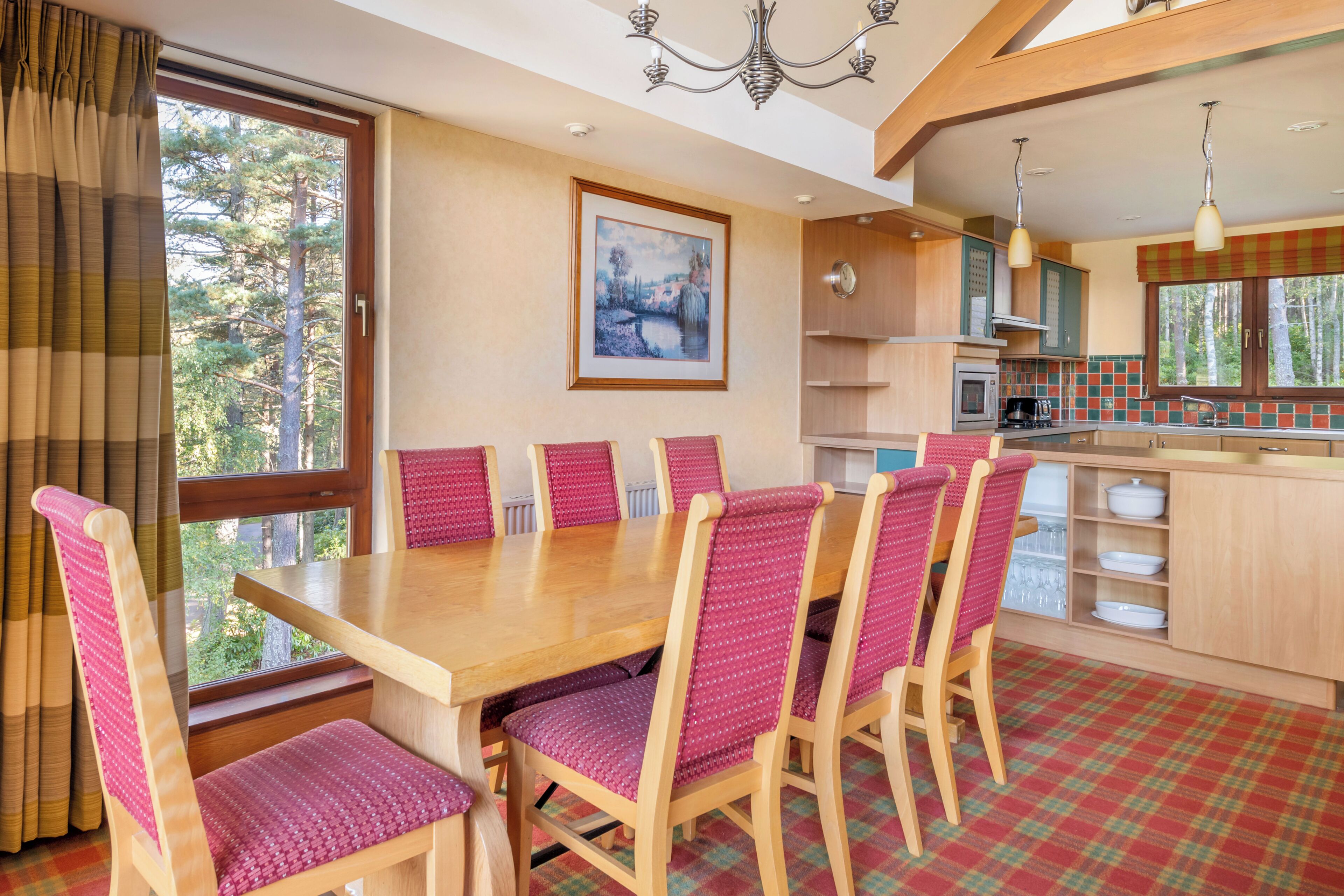 A dining area with a wooden table and chairs, a kitchen with a window, and a painting on the wall.