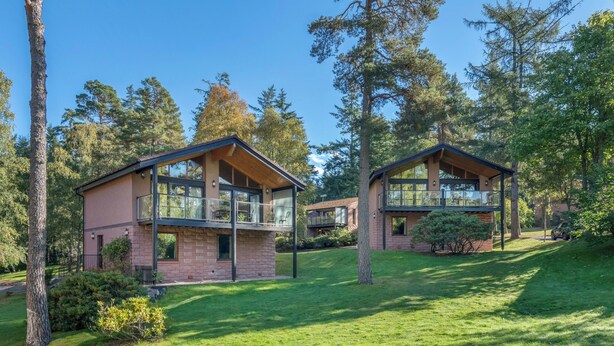 Twee moderne huizen met grote glasramen, omgeven door een verzorgd gazon en hoge bomen.