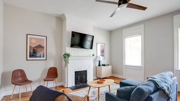 A living room with a fireplace, a television mounted above it, and a ceiling fan. There are two chairs and a sofa. - Walk to VCU & Allianz Amphitheater  Cozy & Clean (Richmond)