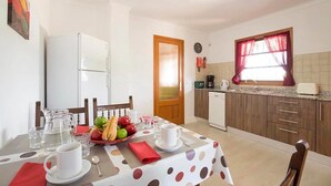 A dining area with a table set for two, a refrigerator, a coffee maker, and a window with curtains. - Villa Roqueta (Pollença)