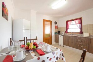 A dining area with a table set for two, a refrigerator, a coffee maker, and a window with curtains. - Villa Roqueta (Pollença)
