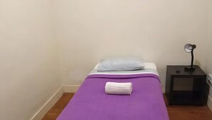 Ein Einzelbett mit lila Bettdecke, einem weißen Kopfkissen und einem aufgerollten weißen Handtuch. Rechts daneben steht ein kleiner Nachttisch mit einer Lampe.
