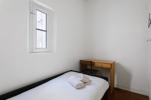 Ein kleines Zimmer mit einem Einzelbett, einem Holztisch, einem Fenster und einem Handtuch auf dem Bett.