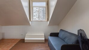 Ein Zimmer mit schräg verlaufender Decke, einem Fenster mit Blick auf Gebäude, einer Couch, einer Bank und einem Fliesenboden.