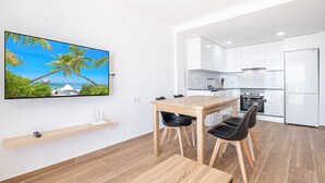 Eine moderne Küche mit weißen Schränken, einem Holzespeisetisch mit schwarzen Stühlen und einem an der Wand befestigten Fernseher, der eine tropische Szenerie zeigt.