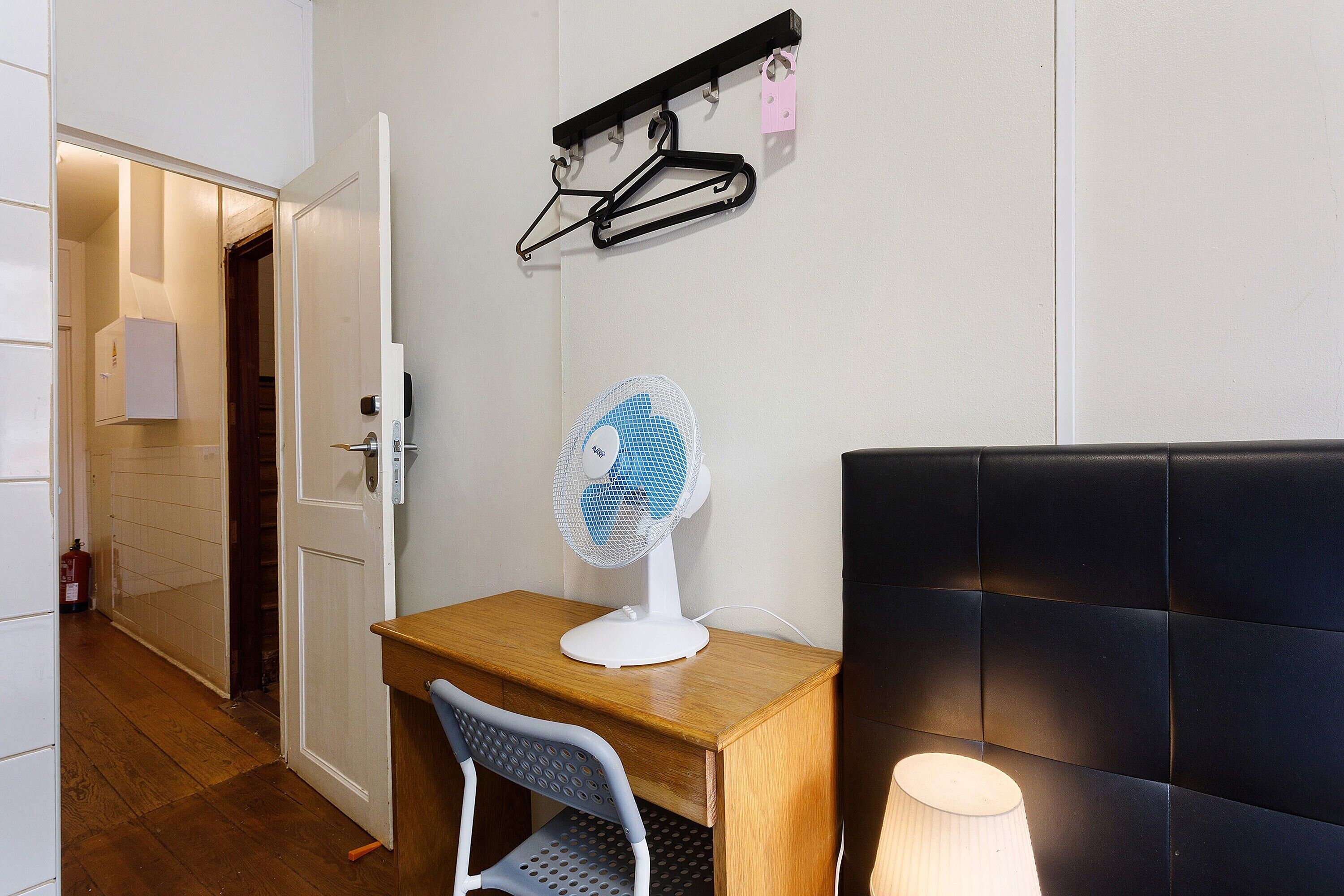 Ein Zimmer mit Schreibtisch aus Holz, einem weißen Stuhl, einem Standventilator, einem schwarzen Kopfteil und einem an der Wand befestigten Kleiderhaken mit Kleiderbügeln.