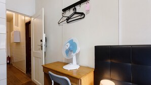 Et værelse med skrivebord i træ, hvid stol, en ventilator, sort sengegavl og et vægmonteret knagered med knager.