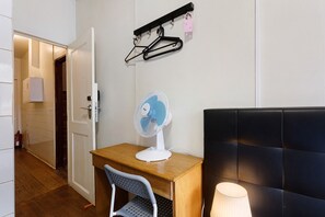 Une pièce avec un bureau en bois, une chaise blanche, un ventilateur sur pied, une tête de lit noire et un porte-manteau mural avec des cintres.