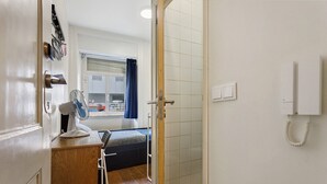 Et soveværelse med en seng, et skrivebord med stol, en ventilator, et vindue med gardiner og en telefon på væggen.