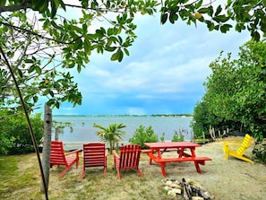 A beachside view with a clear sky, a calm sea, and a row of red and yellow chairs and a picnic table. - Cozy studio in Caye Caulker with AC and rooftop patio (Caye caulker)