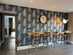 A room with a peacock-patterned wallpaper, a wooden bar table with metal chairs, a wall-mounted clock, and recessed ceiling lighting.