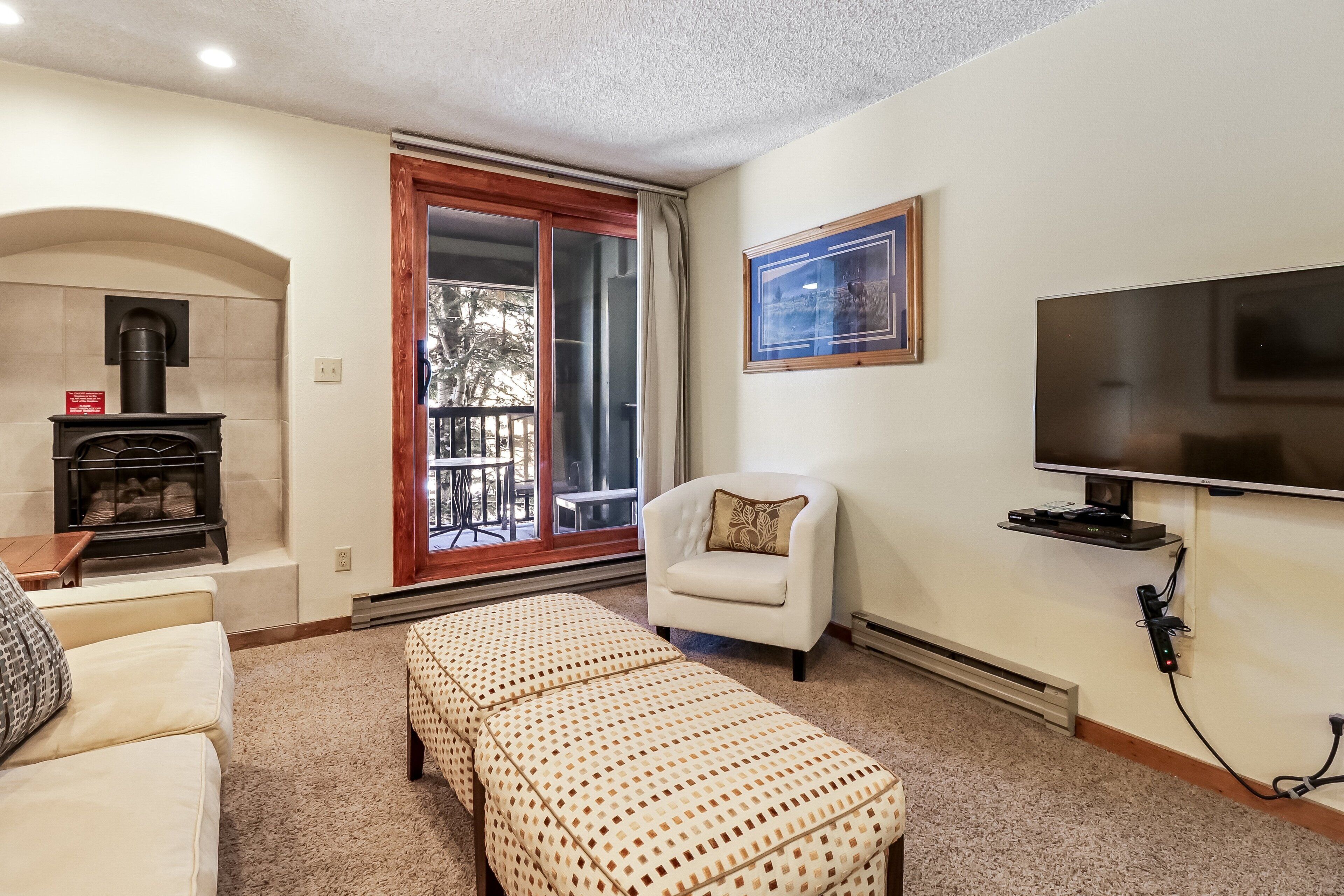 A living room with a fireplace, a television, a sofa, an ottoman, and a sliding glass door leading to an outdoor area.
