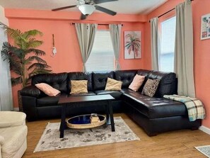 A living room with a black leather sofa, a coffee table, a plant, and a ceiling fan. - Coastal Oasis In Surfside Beach 3 Bedroom Home (Freeport)