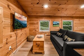 A wooden interior with a black sofa, a wooden coffee table, and two windows with views of greenery. - Shamrock Valley 1 Bedroom Cabin (Sevierville)