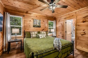 Un dormitorio de cabaña de madera con una cama, dos mesitas de noche, un ventilador de techo y una ventana con vistas a árboles.