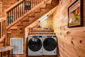A wooden cabin interior with a staircase, two front-loading washers, a small table with a magazine, and a framed poster on the wall. - Shamrock Valley 1 Bedroom Cabin (Sevierville)