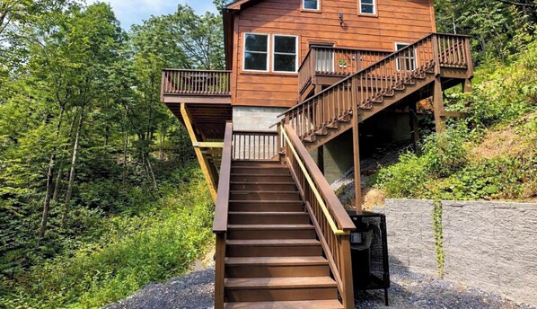 A wooden cabin with a staircase leading to a balcony, surrounded by dense forest. - Shamrock Valley 1 Bedroom Cabin (Sevierville)