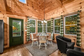 Interior de una cabaña de madera con una mesa de comedor, sillas, un refrigerador y ventanas grandes con cortinas.