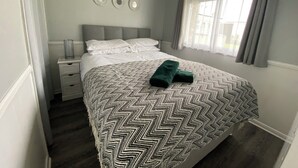 A neatly made bed with a patterned bedspread, a bedside table with a lamp, and a window with curtains. - Chalet 57 (Great Yarmouth)