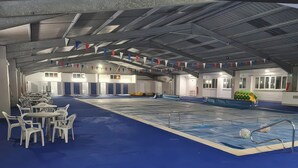 Piscina interna com piso azul, rodeada por cadeiras e mesas de plástico branco, e teto com vigas de metal e bandeiras triangulares.