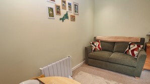 A dining area with a table set for two, a radiator, a green sofa with patterned cushions, and framed pictures on the wall. - Mini Mena (St Austell)