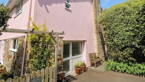 A pink building with a stone chimney, a wooden pergola, a wooden bench, and a welcome sign. - Mini Mena (St Austell)