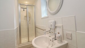 A bathroom with a glass shower enclosure, a white sink with a marble soap dispenser, and a wall-mounted oval mirror. - Mini Mena (St Austell)