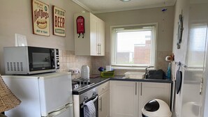 Cocina compacta con microondas, estufa, horno y pileta. Hay placares, una ventana con persianas y un bote de basura.