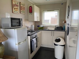 Cozinha compacta com micro-ondas, fogão, forno e pia. Há armários, uma janela com persianas e uma lixeira.