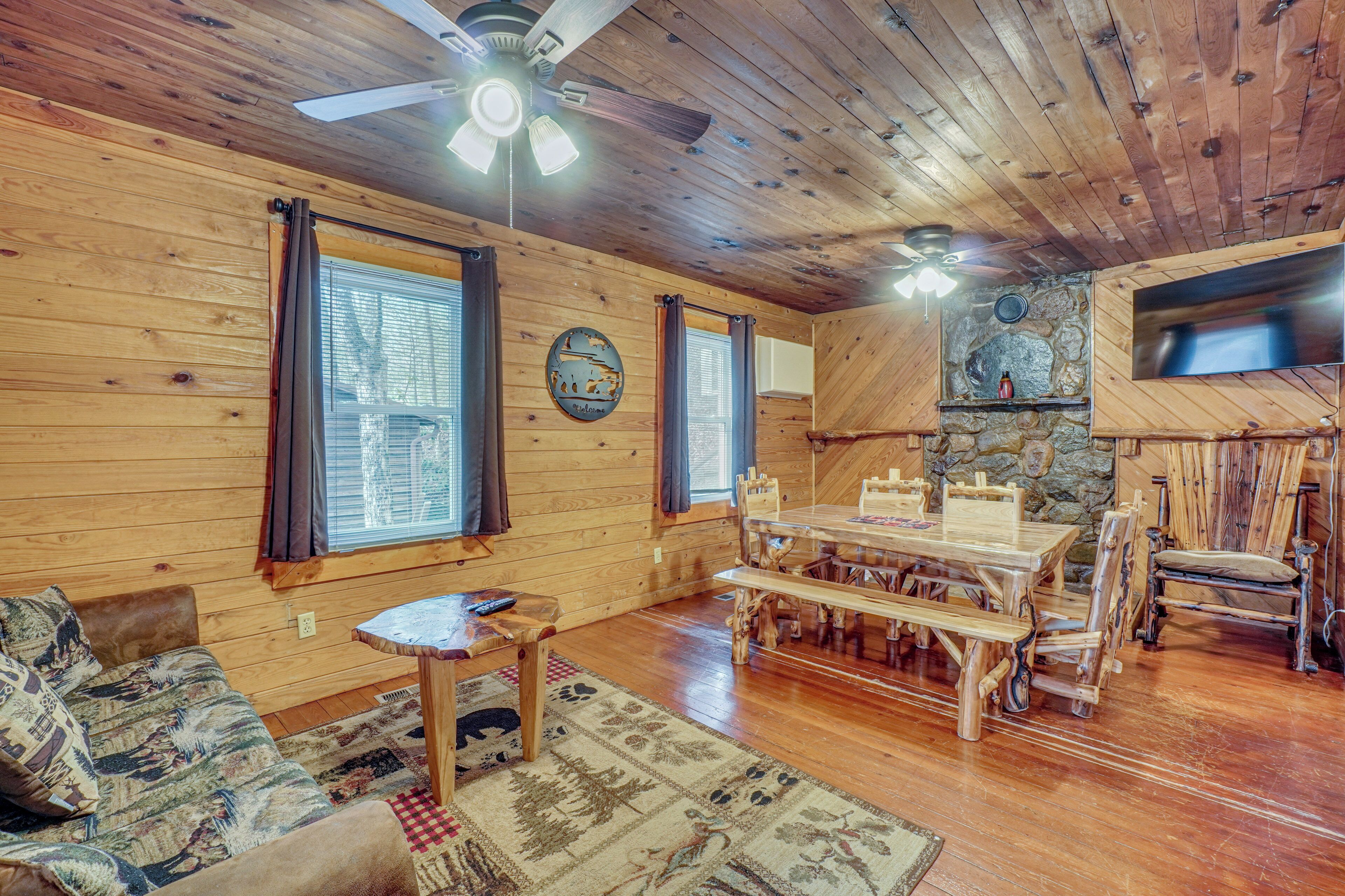 A wooden interior with a ceiling fan, a stone wall, a dining table with benches, and a sofa.