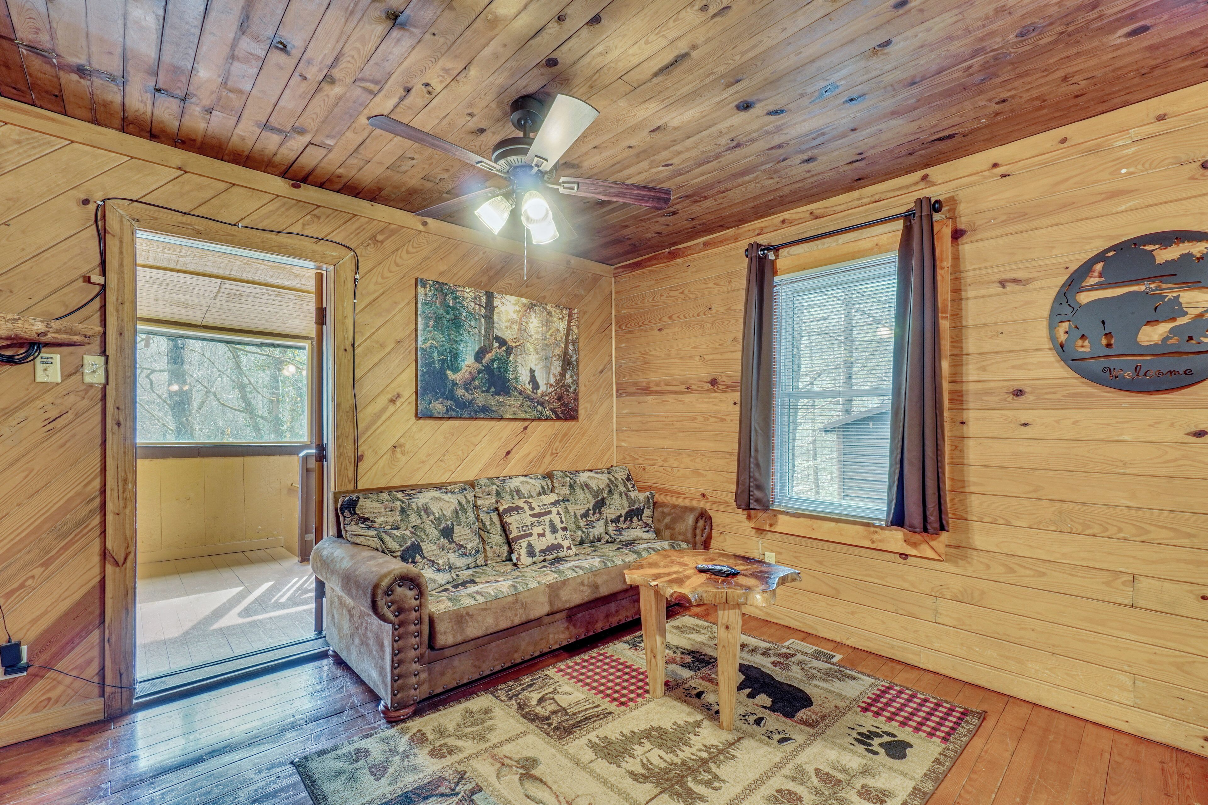 A wooden cabin interior with a sofa, a ceiling fan, a painting, a window, and a welcome sign.