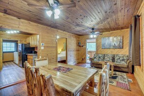 Interior de uma cabana de madeira com uma mesa de jantar, cadeiras, um sofá e um ventilador de teto.