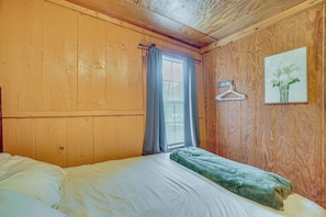 A wooden-paneled room with a bed, a window with curtains, a wall-mounted hanger, and a framed picture.
