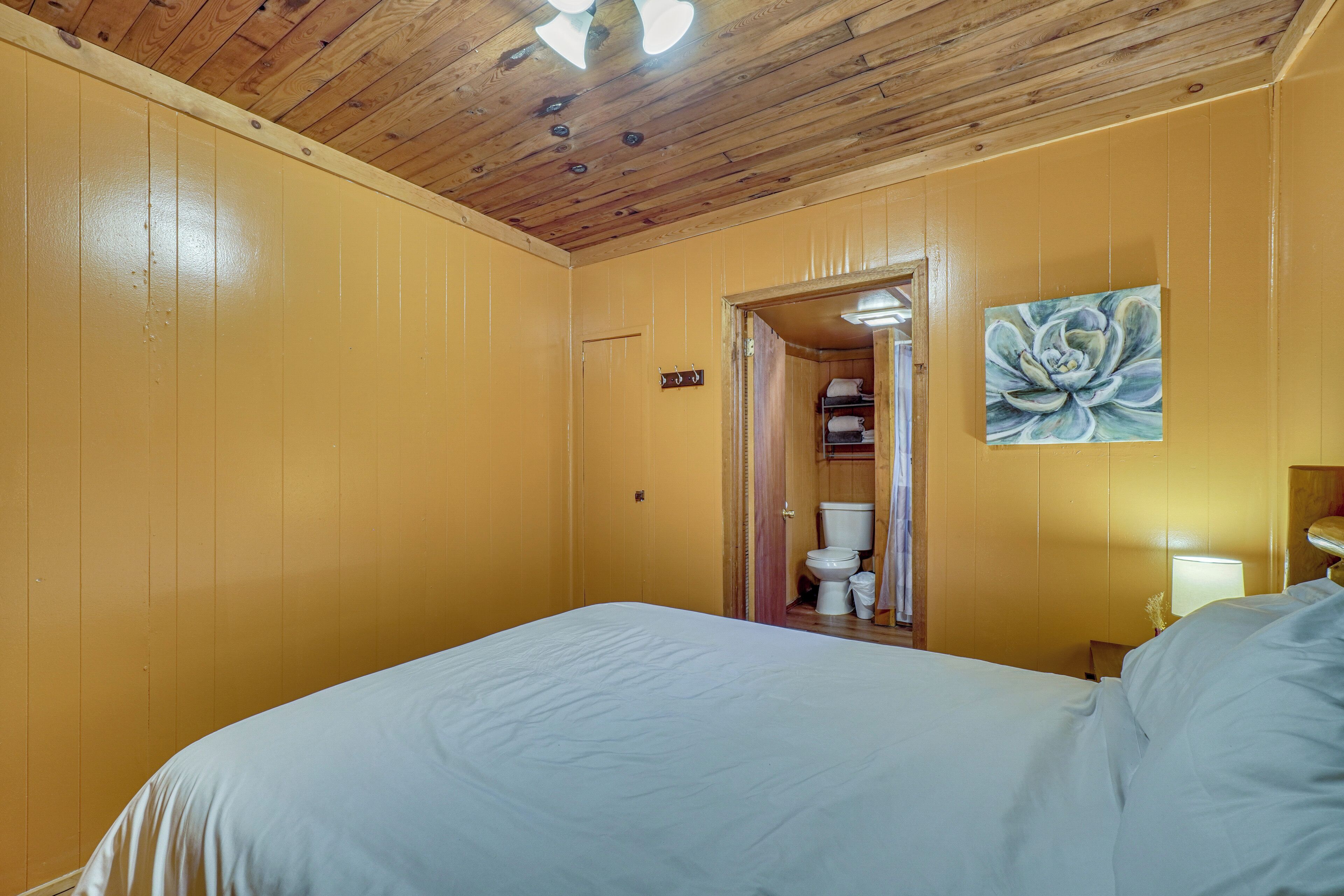 A room with a wooden ceiling, a bed with white bedding, a painting of a flower on the wall, and a door leading to a bathroom.