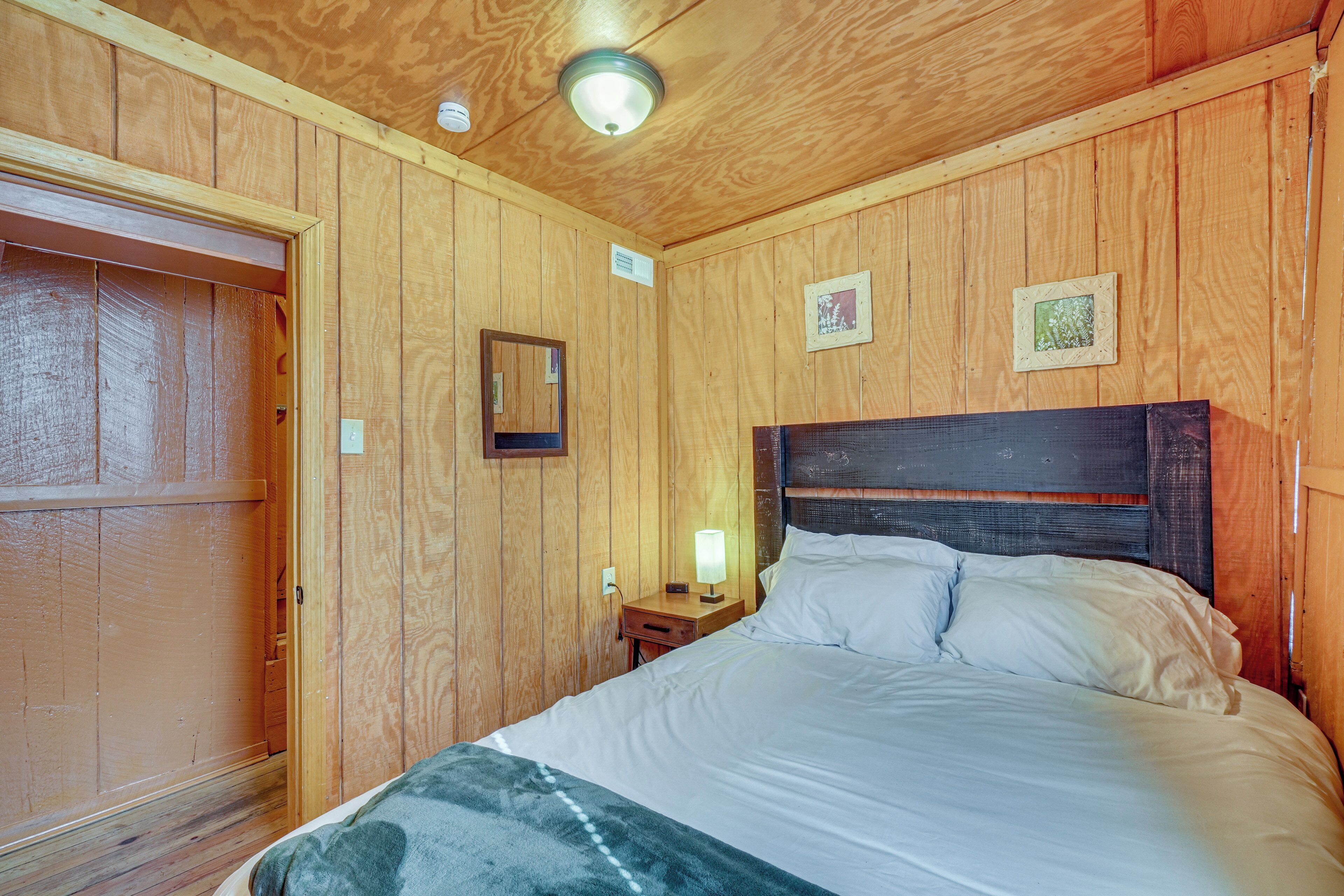 A wooden-paneled room with a bed, a nightstand, a wall-mounted light, and a door.