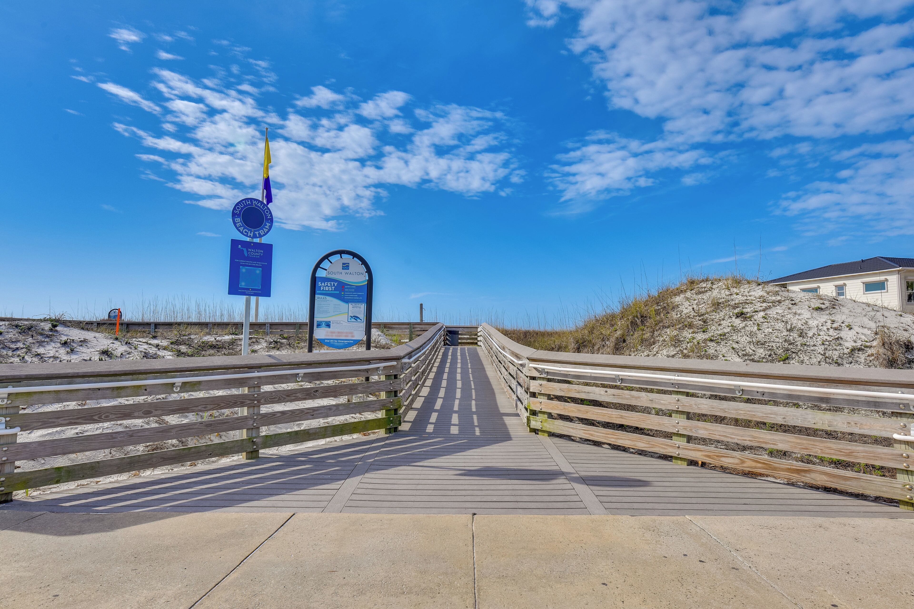 Ein Promenadenweg führt zu einem Strand mit blauem Himmel und einer Flagge.