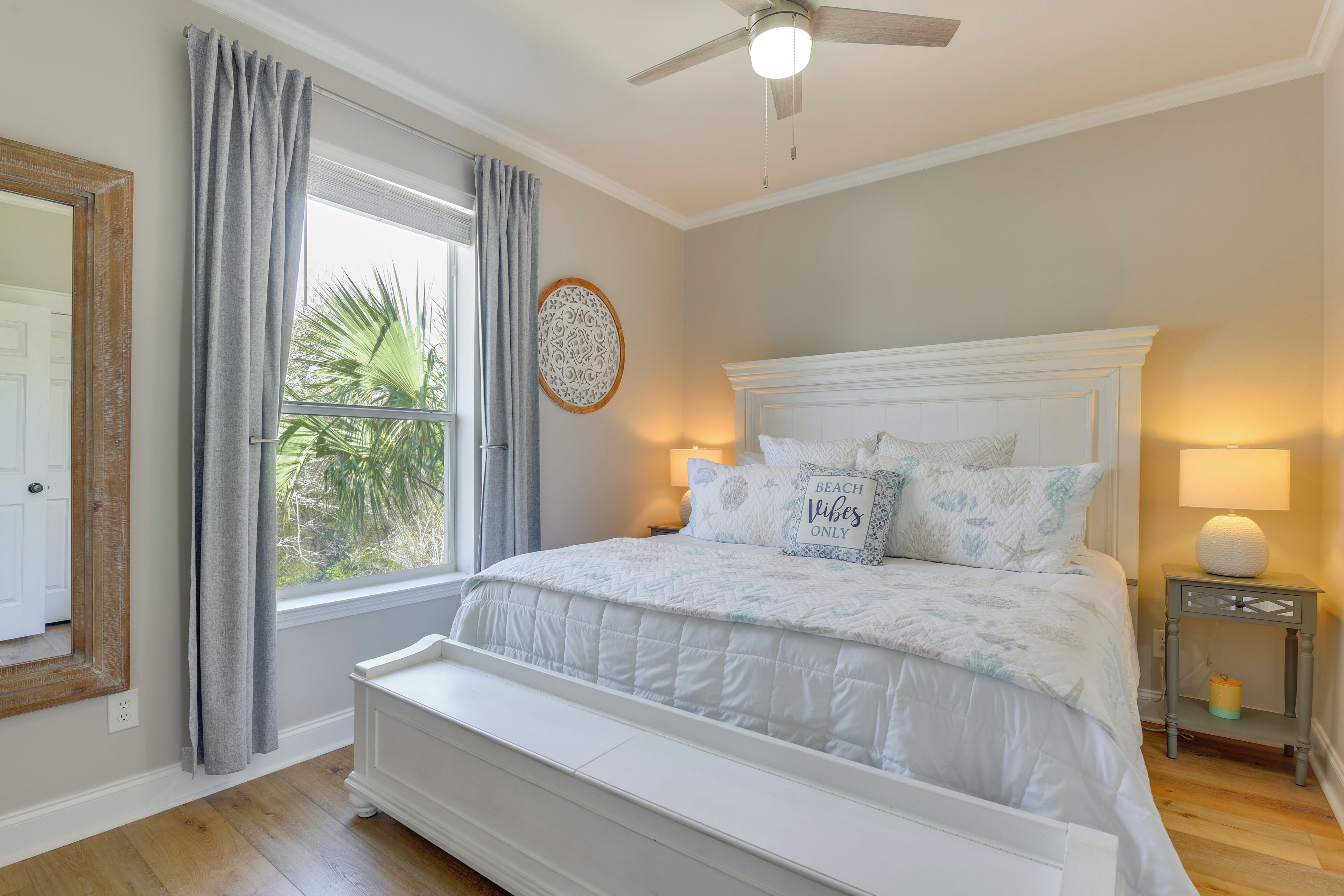 Ein Schlafzimmer mit einem großen Bett, einem Deckenventilator, einem Fenster mit Vorhängen, einem Nachttisch mit Lampe und einer Wanddekoration.