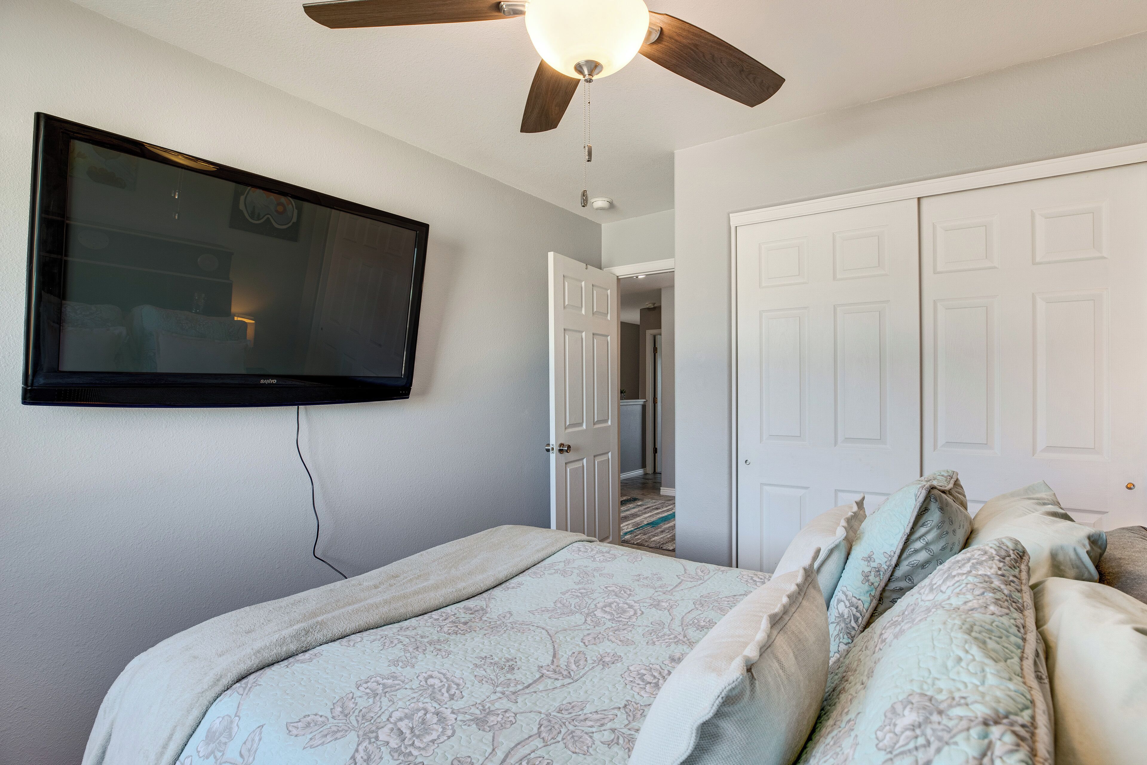 A bedroom with a bed, a ceiling fan, a TV mounted on the wall, and a door leading to another room.