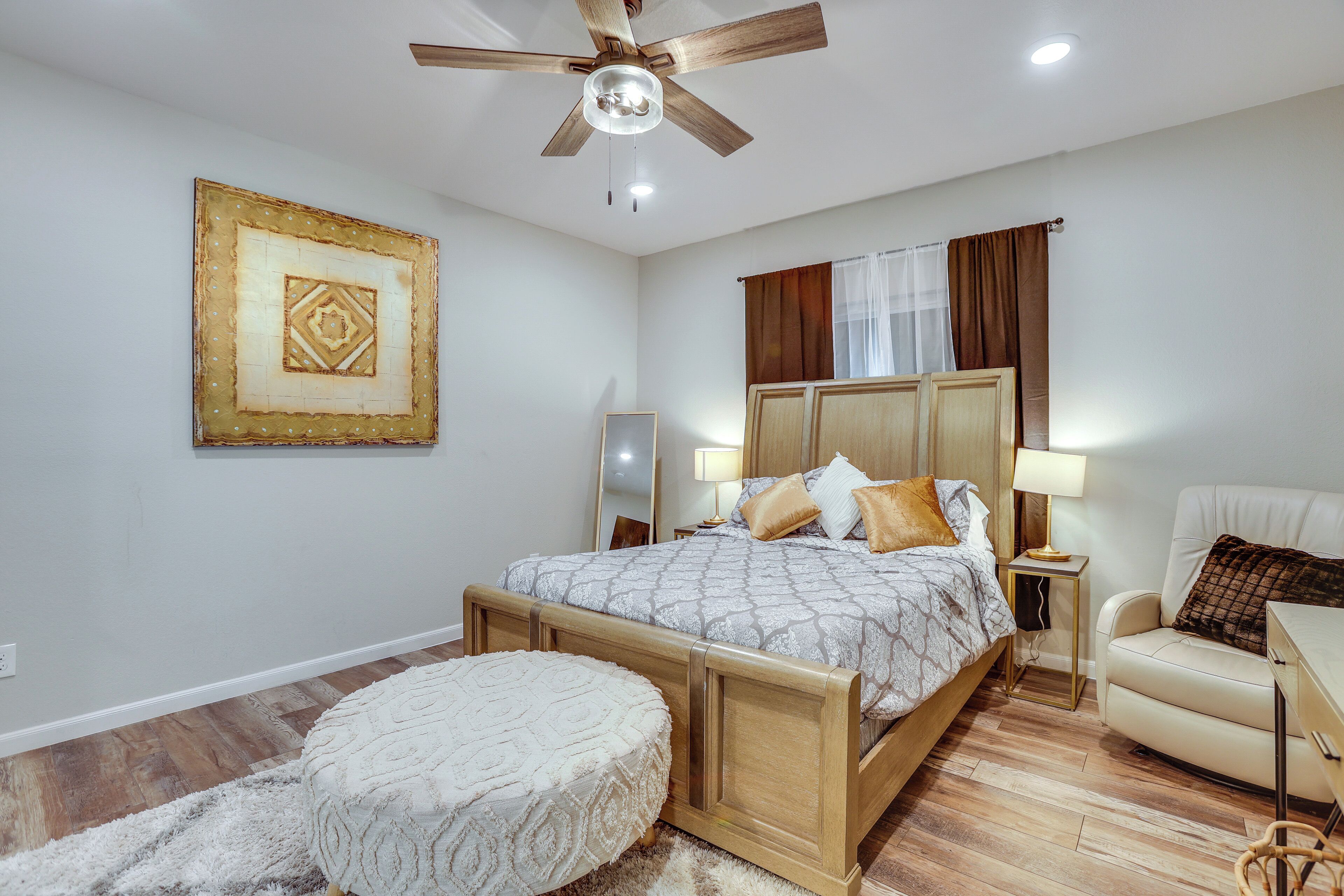 A bedroom with a wooden bed, a round ottoman, a ceiling fan, a bedside table with a lamp, and a framed picture on the wall.