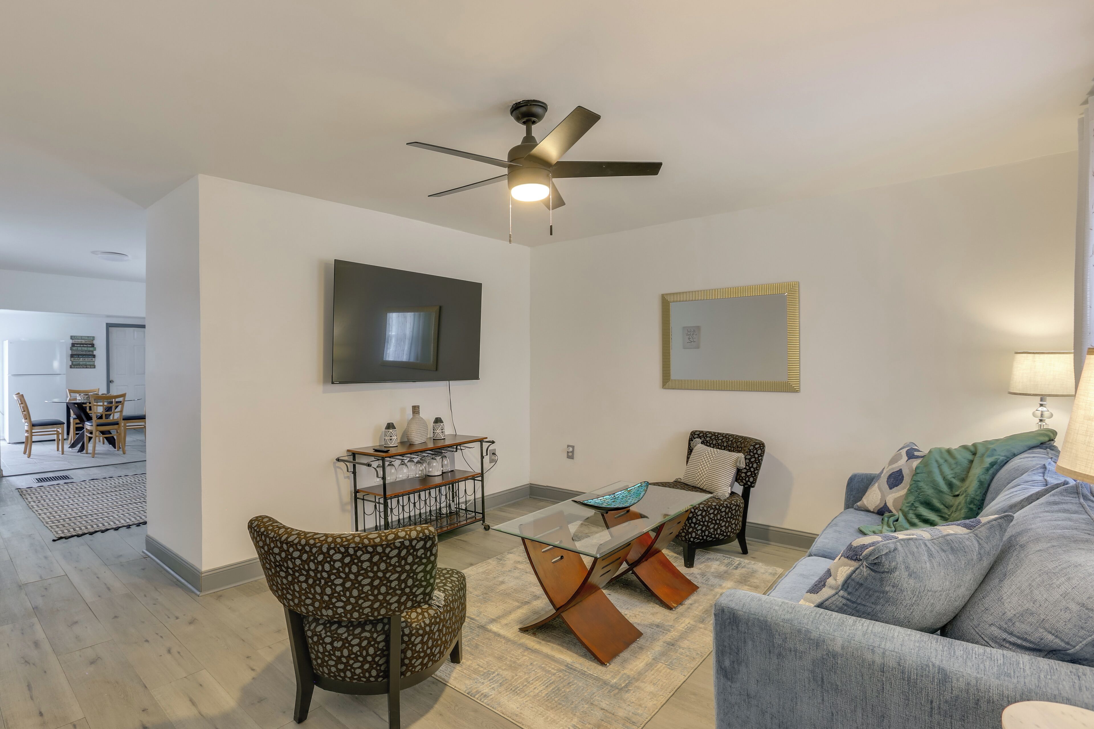 A living room with a ceiling fan, a glass coffee table, patterned chairs, a sofa, and a TV mounted on the wall.