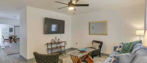 A living room with a ceiling fan, a glass coffee table, patterned chairs, a sofa, and a TV mounted on the wall.