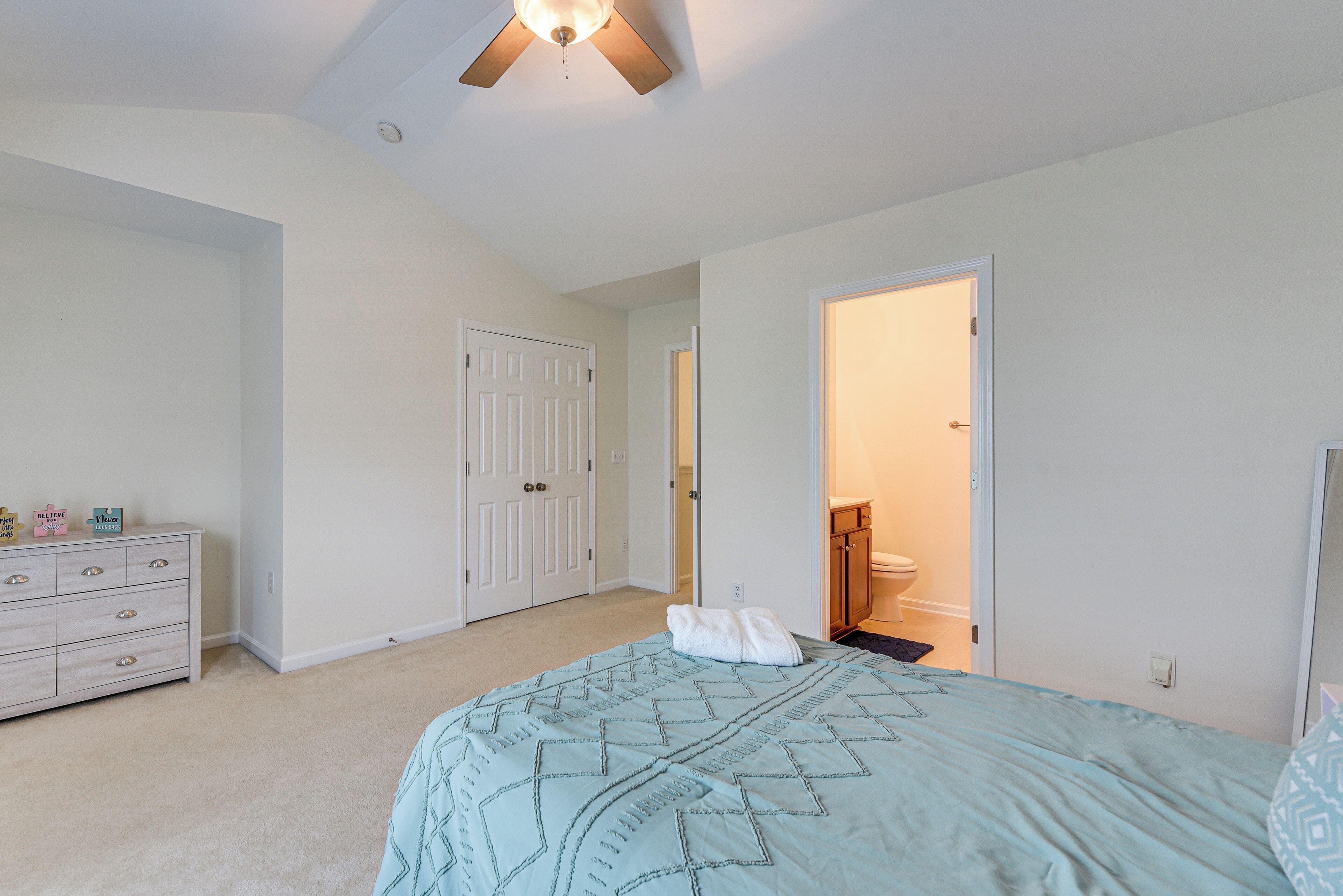 A bedroom with a bed, a dresser, a ceiling fan, and a door leading to a bathroom.
