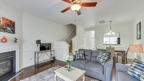 A living room with a fireplace, a ceiling fan, a sofa set, and a dining area with a chandelier. - Dog-friendly Townhome w/ Patio in Charlotte Area! (Matthews)